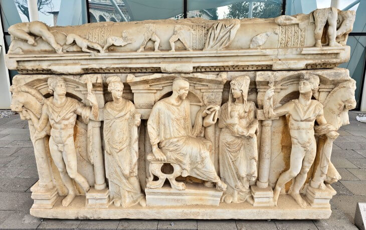 İznik Müzesi açık hava bölümünde sergilenen Roma dönemine ait yüksek kabartmalı mermer lahit; mitolojik figürler, kadın-erkek betimleri ve sahne dekorasyonlarıyla işlenmiş anıtsal mezar örneği.